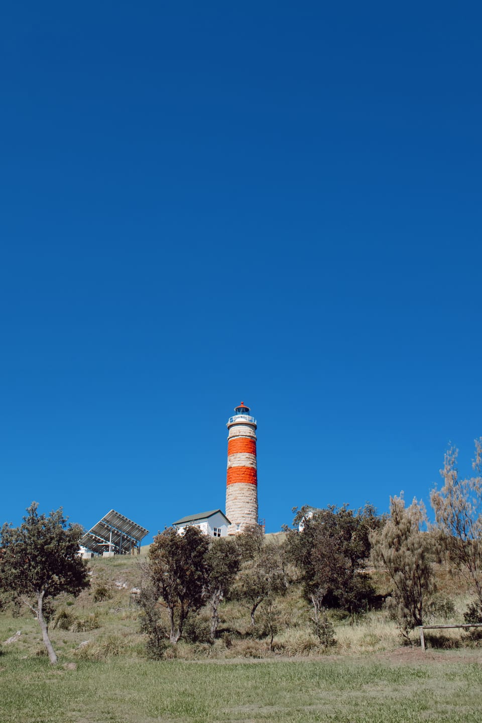 Red-and-white striped sandstone lighthouse on a grassy headland under a clear blue sky, with a solar panel array and low scrub in the foreground