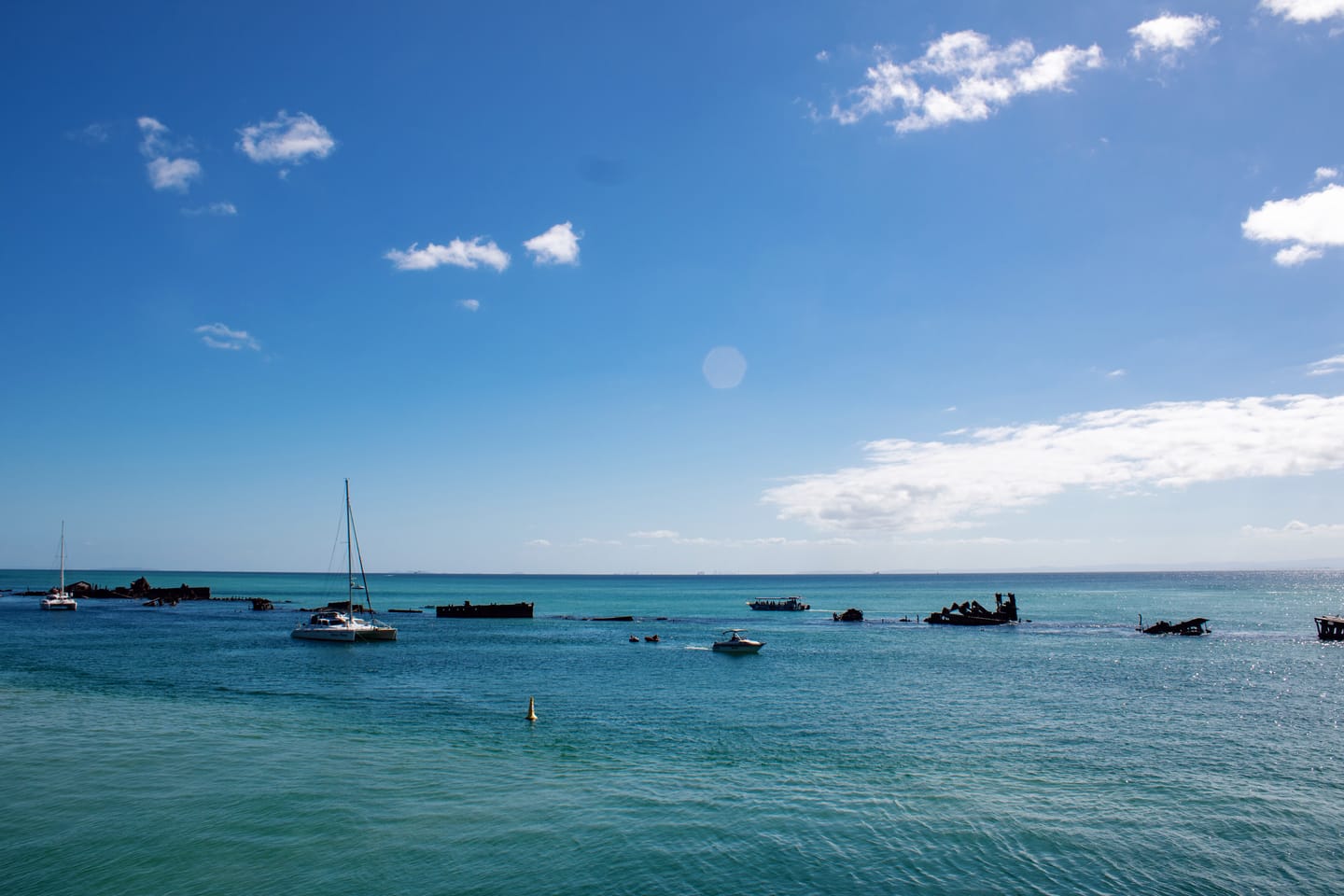 A yacht and small boats among partly submerged, rusted shipwrecks on a calm turquoise bay under scattered clouds