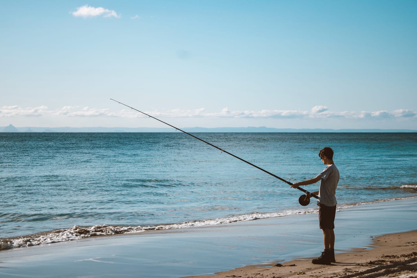 A young man in a grey t-shirt casting a long fishing rod from the water's edge on a sunlit beach