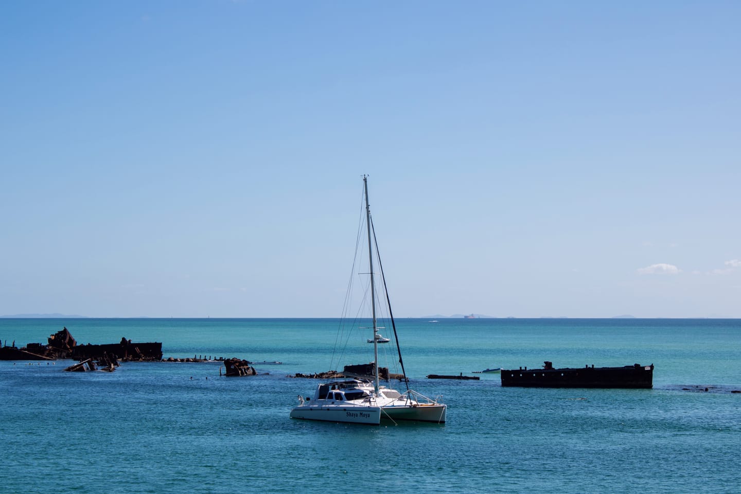 A white catamaran anchored among rusting shipwrecks on flat, pale blue water beneath a hazy sky