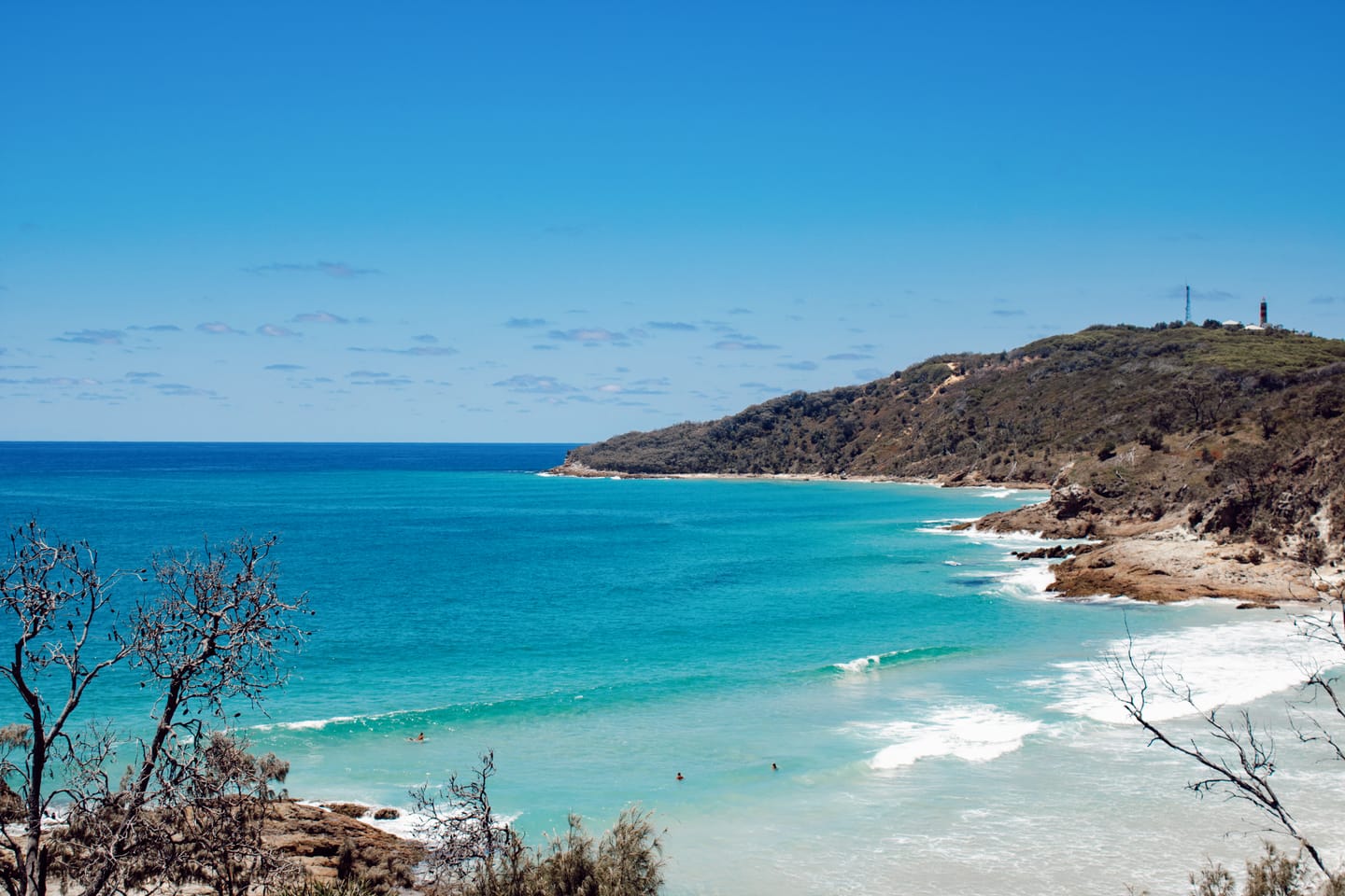 Turquoise surf breaking on a rocky headland below a lighthouse, seen through casuarina branches from a high vantage point