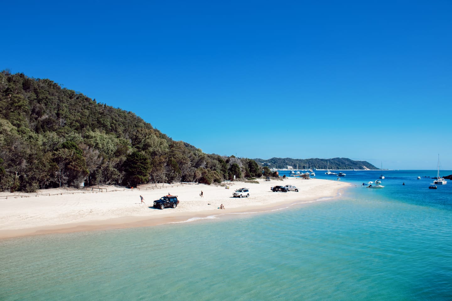 A white-sand beach backed by a forested hill, with four-wheel drives parked on the sand and boats moored in turquoise water